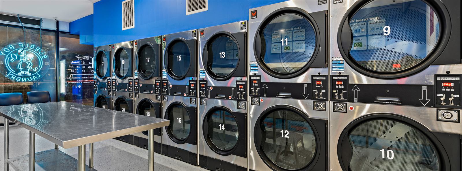 Inside the shop: a bank of numbered stacked dryers along a blue wall, with a long steel folding table in front