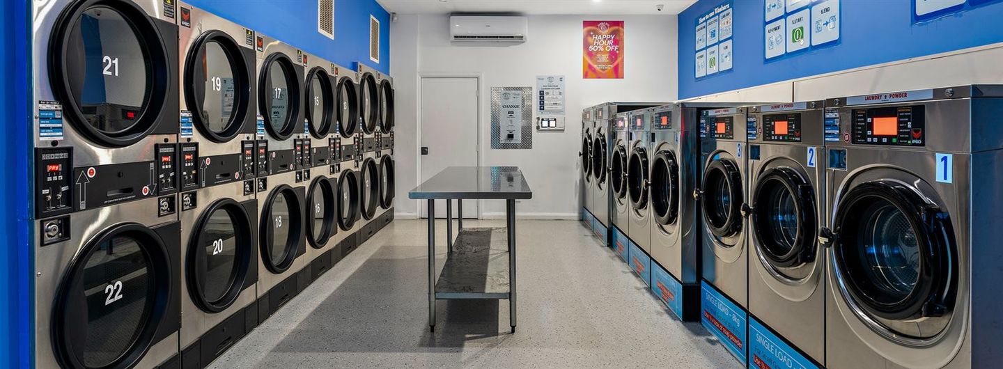 Inside Bubble Boy Laundry: eight front-load washers down the right wall, stacked dryers numbered 9 to 22 on the left, a long steel folding table in the middle and the change machine on the back wall