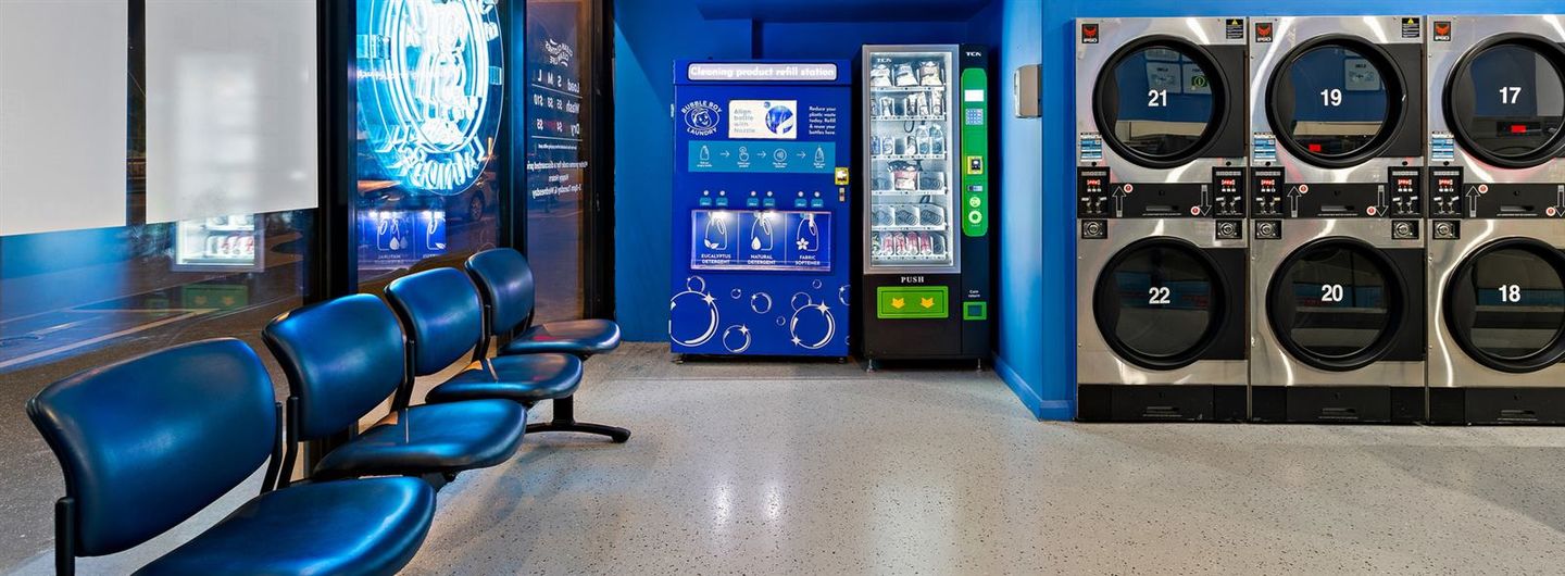 The waiting area: four blue chairs by the window, the detergent refill station and vending machine beside them, dryers to the right