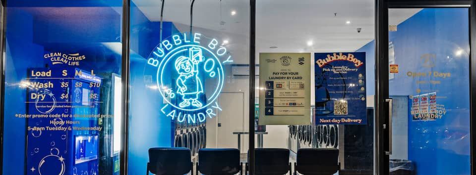 The Bubble Boy Laundry shopfront at night &mdash; blue neon Bubble Boy sign glowing in the window, with the washers and dryers visible inside